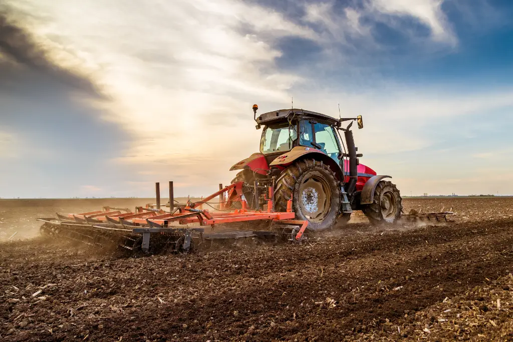 Massey Ferguson tractor cultivating a large farm field at sunset with dust rising behind it