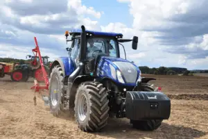 A blue New Holland tractor driving across a dry plowed field with a mounted red agricultural implement.