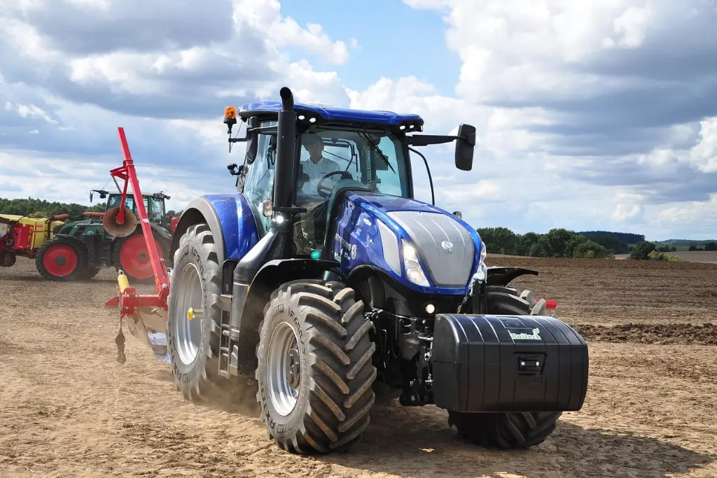 A blue New Holland tractor driving across a dry plowed field with a mounted red agricultural implement.