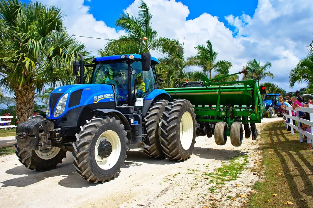 Blue New Holland tractor pulling a green agricultural seeder down a palm-lined path with spectators watching from the side.